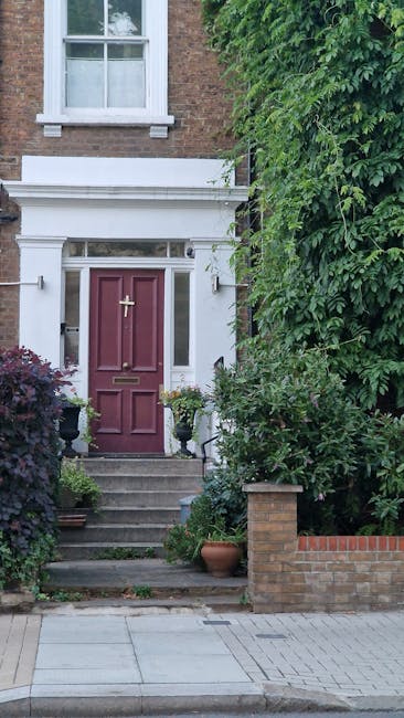 A residential garden entrance featuring a deep red wooden front door with a brass handle and mail slot, accessed via a small set of concrete steps adorned with potted plants and flower pots. Flanking