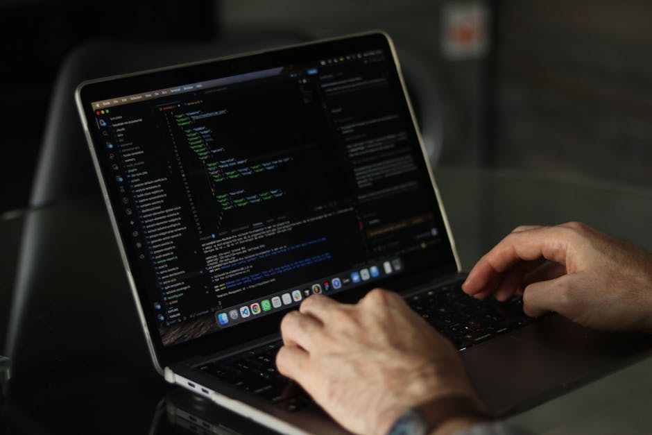 A person working on a laptop with a dark-themed coding interface displayed on the screen, sitting at a black table. The individual’s hands are visible, with one hand poised above the keyboard, possibl