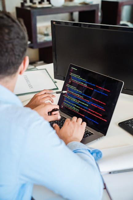 A person working on a laptop with a dark-themed coding interface displayed on the screen, sitting at a black table. The individual’s hands are visible, with one hand poised above the keyboard, possibl